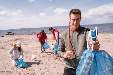 Bulanık gönüllü grubunun yanında elinde çöp torbası tutan mutlu bir adam kumda çöp topluyor. 