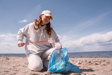 Kapüşonlu mutlu kadın çöp torbası tutuyor ve kumda çöp topluyor. 