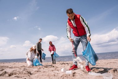 Genç adam çöp torbası tutuyor ve gönüllülerin yakınındaki kumda çöp topluyor. 