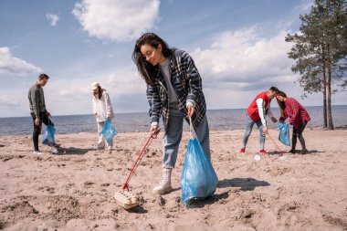 smiling young woman holding trash bag and collecting rubbish on sand near group of volunteers  clipart