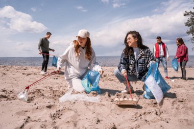 smiling young women holding trash bags and collecting rubbish with grabbers on sand near group of volunteers  clipart