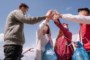 Tıbbi maskeli genç kadınlar ve erkekler çöp torbalarını tutarken ve el ele tutuşurken çöp konseptini seçiyorlar. 