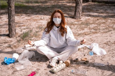 Tıbbi maskeli genç bir kadın yerdeki çöplerin yanında meditasyon yapıyor.