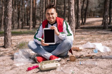 Bacak bacak üstüne atmış mutlu bir adam oturmuş elinde tabletle yerde çöp kutusunun yanında boş bir ekran tutuyor.