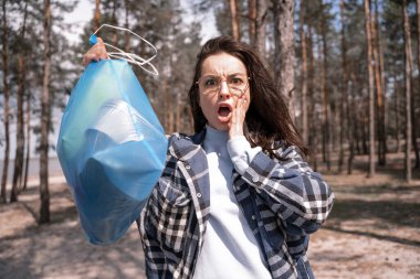Şok olmuş genç bir kadın ormanda mavi çöp torbası tutuyor. 