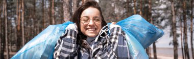 Ormanda mavi çöp torbaları taşıyan gözlüklü neşeli genç bir kadın.