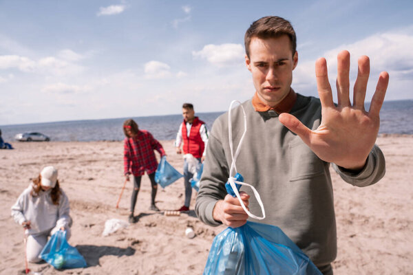 young man showing stop gesture near blurred group of volunteers picking up trash, ecology concept 