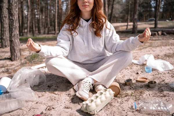 足を交差させ地面のゴミの近くで瞑想している若い女性のクロップドビュー — ストック写真