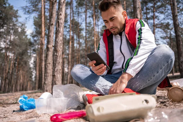 Bacak bacak üstüne atmış bir adam oturuyor ve çöp tenekesinin yanında akıllı telefon kullanıyor.