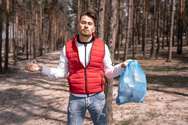 Молодой человек держит синий мешок для мусора и пластиковую бутылку в лесу 