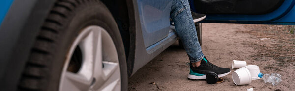cropped view of man standing on ground near car and trash, ecology concept, banner