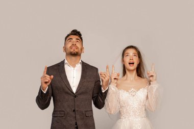 astonished bride and groom looking up and pointing with fingers isolated on grey