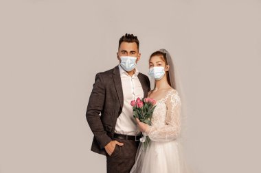 man in medical mask standing with hand in pocket near bride with tulips isolated on grey