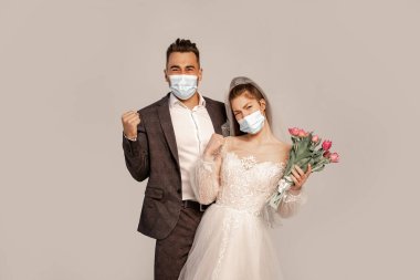 excited newlyweds in protective masks showing yeah gesture isolated on grey