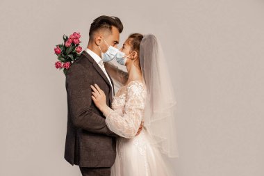 side view of bride and groom in protective masks standing face to face isolated on grey