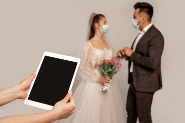 woman holding digital tablet with blank screen near blurred newlyweds in medical masks isolated on grey