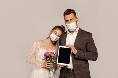 bride and groom in medical masks, with digital tablet and tulips isolated on grey