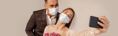 bride in medical mask taking selfie with groom isolated on grey, banner