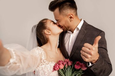 man showing thumb up while kissing bride isolated on grey with lilac shade