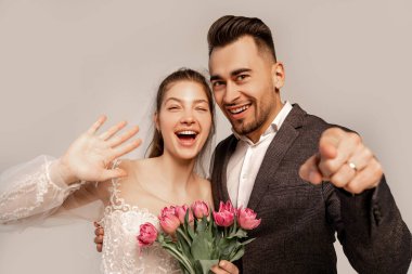 cheerful couple pointing with finger and waving hand isolated on grey with lilac shade