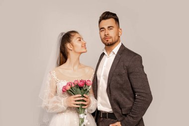 happy bride looking at confident groom standing with hand in pocket isolated on grey
