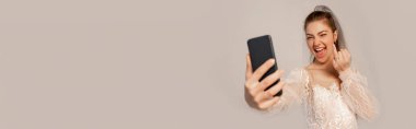 excited bride showing wedding ring while taking selfie isolated on grey, banner