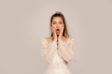 shocked bride touching face while looking at camera isolated on grey with lilac shade