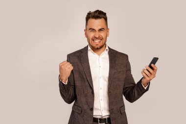 excited man with mobile phone showing win gesture isolated on grey clipart