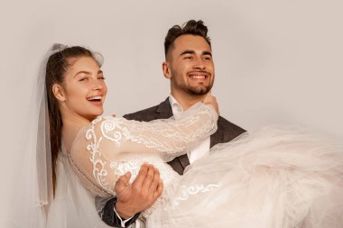 happy man holding cheerful bride in hands isolated on grey