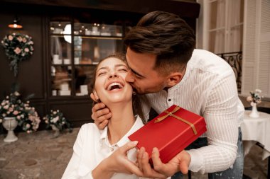 young man kissing laughing woman while presenting gift in cafe