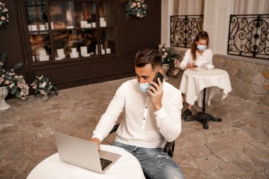 man in medical mask talking on smartphone near laptop and young woman on blurred background