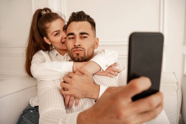 joyful woman biting ear of boyfriend taking selfie on smartphone on blurred foreground
