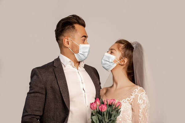 newlyweds in medical masks looking at each other isolated on grey with lilac shade