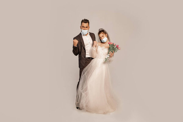 excited newlyweds in medical masks showing triumph gesture isolated on grey with lilac shade