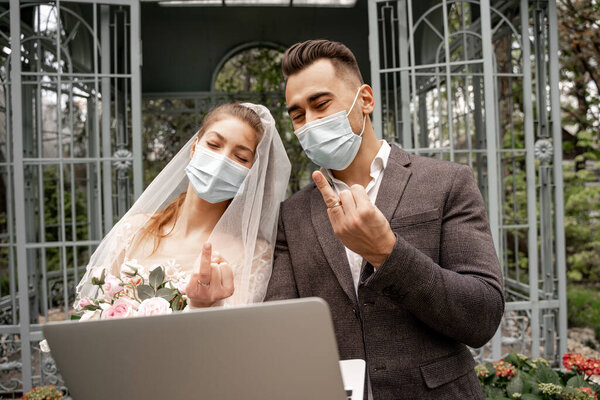 newlyweds in protective masks showing wedding rings during video chat on laptop