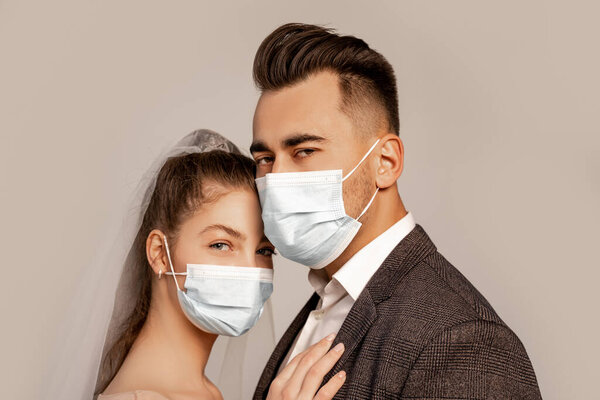 young bride and groom in medical masks looking at camera isolated on grey with lilac shade