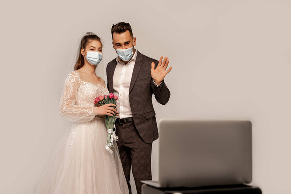 groom in medical mask waving hand during video call on blurred laptop isolated on grey