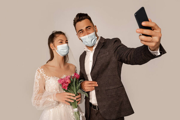 newlyweds in safety masks taking selfie isolated on grey with lilac shade