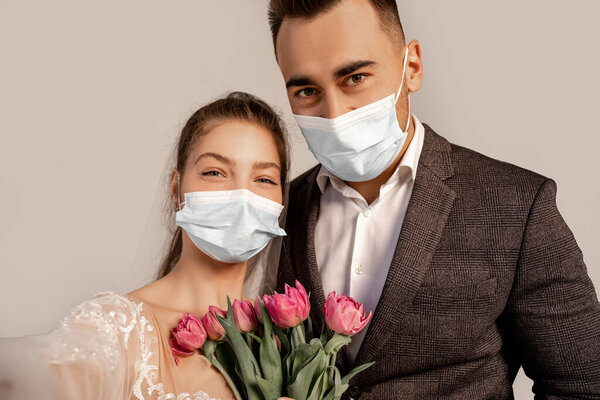 young newlyweds in medical masks looking at camera near fresh tulips isolated on grey