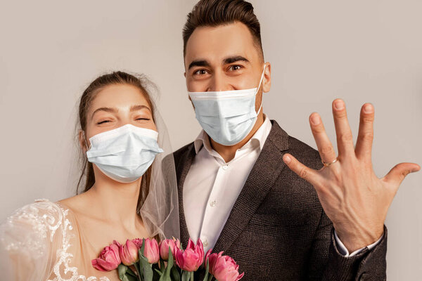 groom in medical mask showing wedding ring near bride with tulips isolated on grey