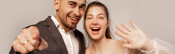 cheerful newlyweds waving hand and pointing with finger isolated on grey, banner