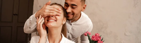 Happy Man Covering Eyes Cheerful Girlfriend Hand While Holding Flowers — Stok fotoğraf