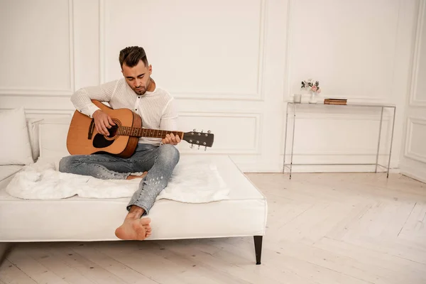 barefoot man in jeans playing acoustic guitar on sofa at home