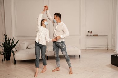 young barefoot couple in jeans and medical masks dancing at home