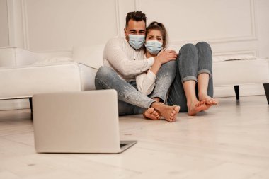 scared couple in medical masks watching horror film on laptop on floor
