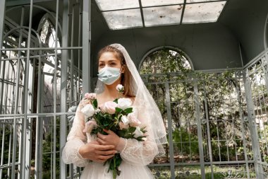 young bride in safety mask standing with wedding bouquet near alcove in park