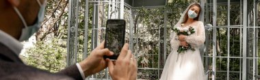 blurred man taking photo of bride in medical mask on smartphone in park, banner