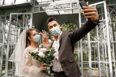 newlyweds in medical masks taking selfie on mobile phone in park