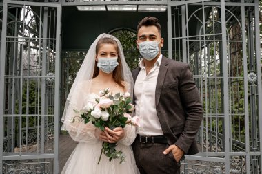 elegant newlyweds in medical masks looking at camera near alcove in park