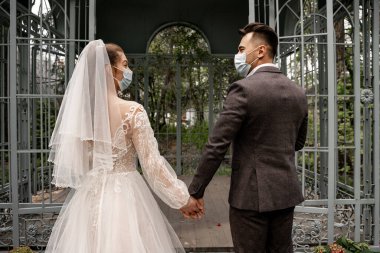 back view of bride and groom in medical masks holding hands near alcove in park